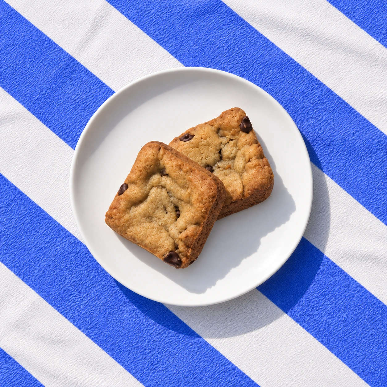 Two chocolate chip cookie bars on a white plate over blue and white striped fabric