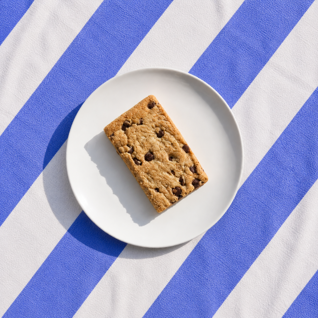 A chocolate chip cookie bar on a white plate over blue and white striped fabric