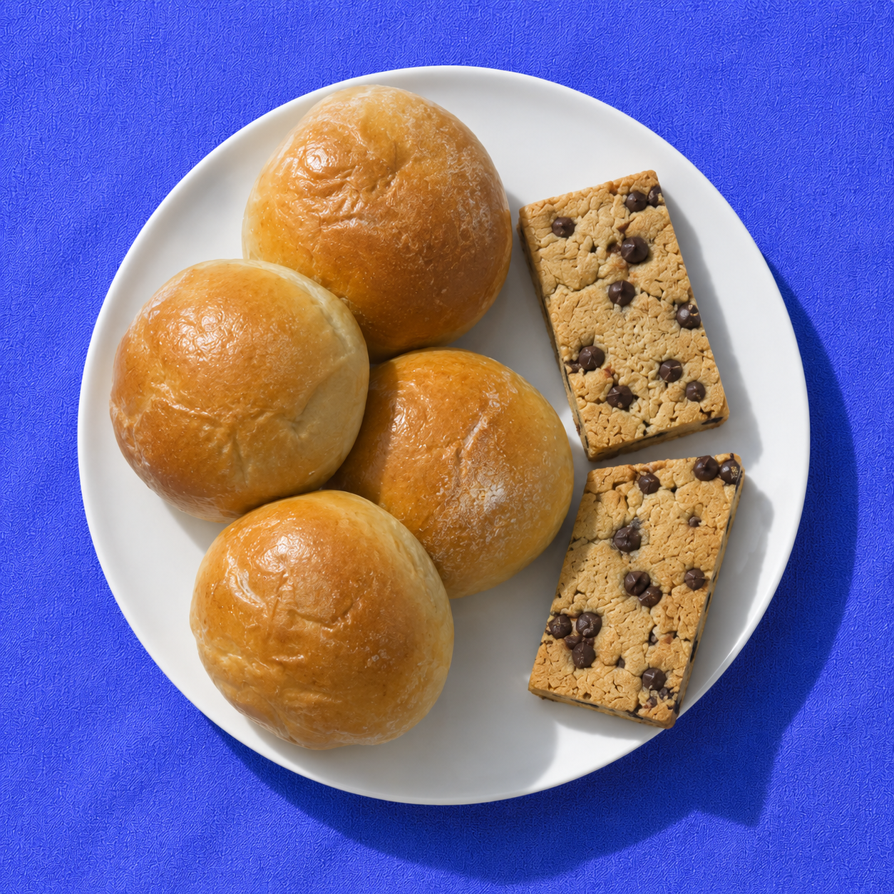 Four beef bierocks and two chocolate chip cookie bars on a white plate over vivid blue fabric
